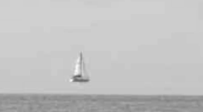 Fotografia de um veleiro no horizonte com efeito de miragem sobre o mar