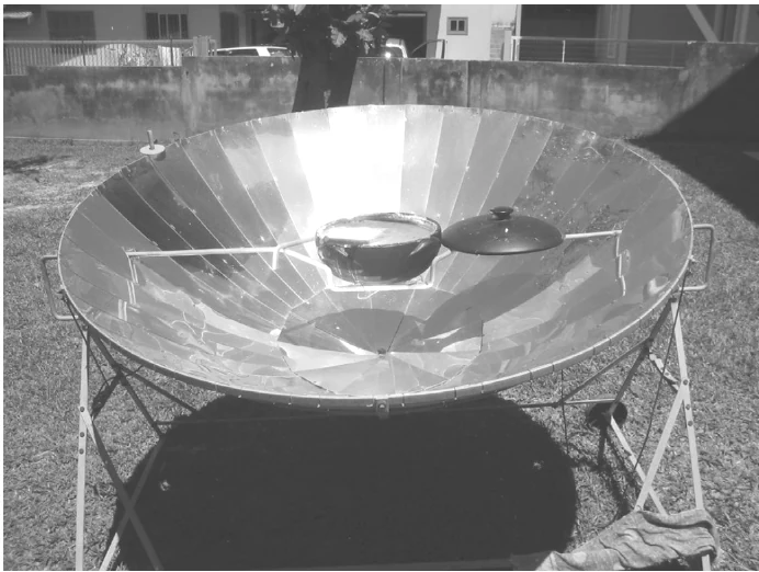 Fotografia de um fogão solar: uma grande superfície refletora parabólica, formada por faixas espelhadas, com uma panela e uma tampa apoiadas sobre ela