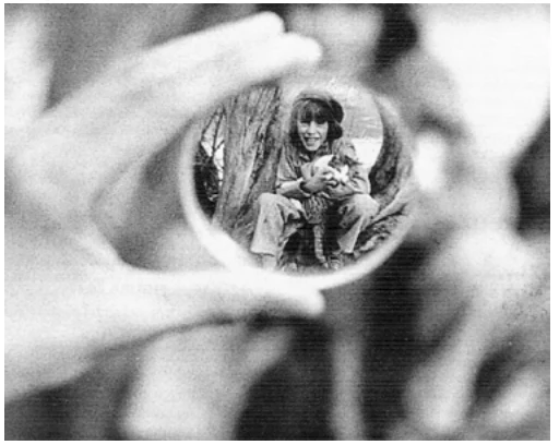 Fotografia em preto e branco: uma mão segura um objeto circular de vidro, e por ele se vê, menor, a imagem de uma menina com um gato no colo