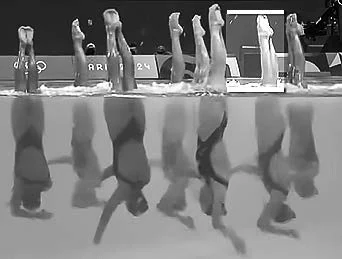 Equipe de nado artístico em posição invertida dentro da piscina, com as pernas para fora da água